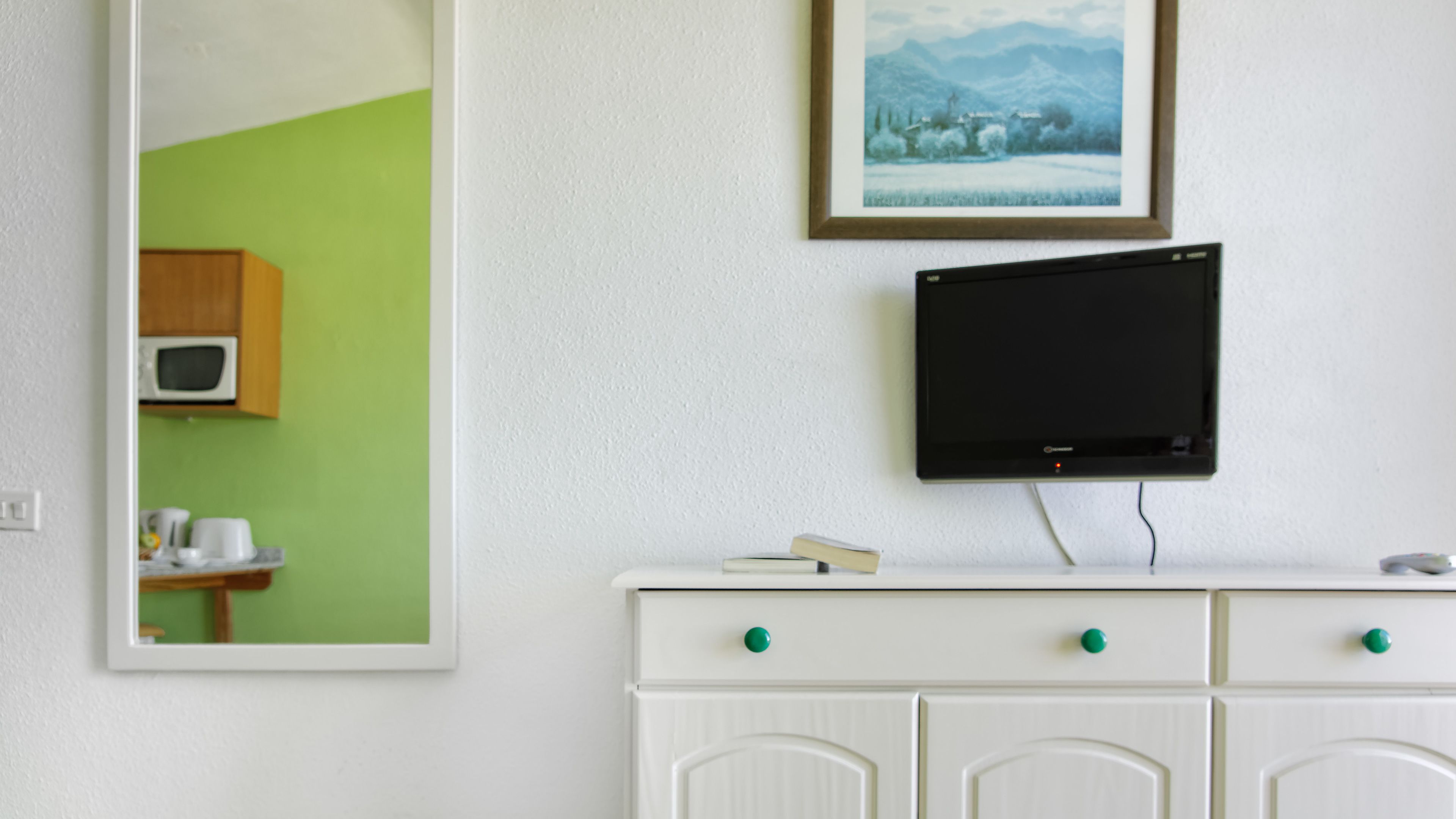 Une pièce avec une commode blanche, un téléviseur à écran plat fixé au mur au-dessus, un mur vert et un tableau encadré placé au-dessus du téléviseur.