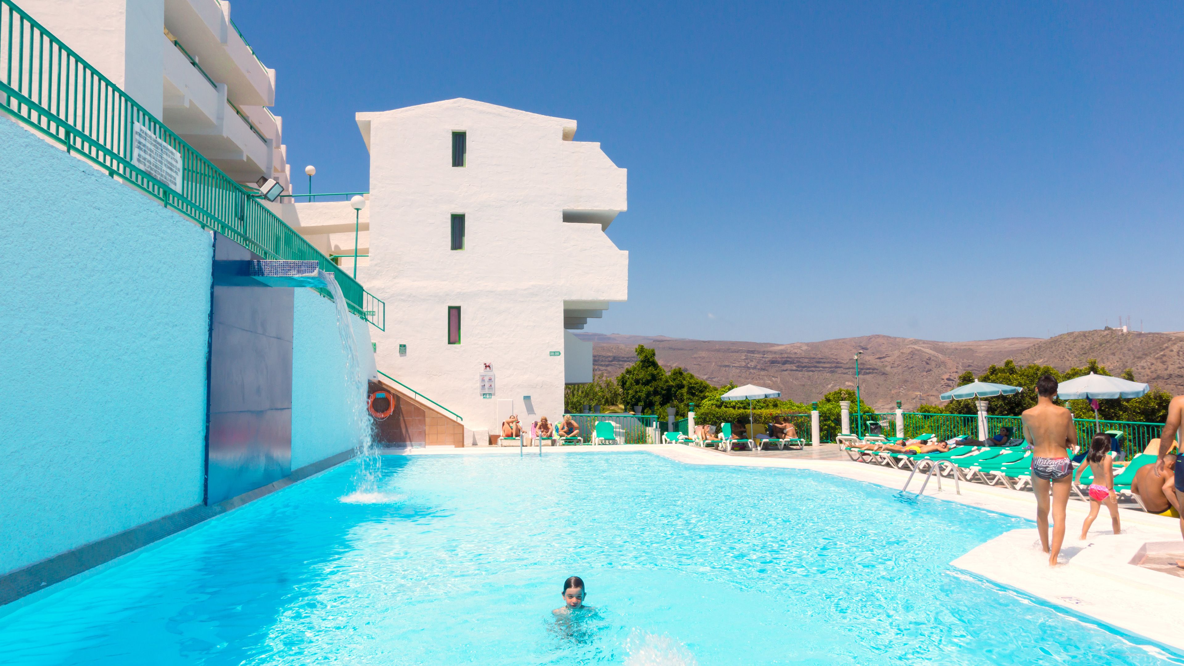 A swimming pool with a clear blue water, a white building with balconies, and a view of hills in the background.