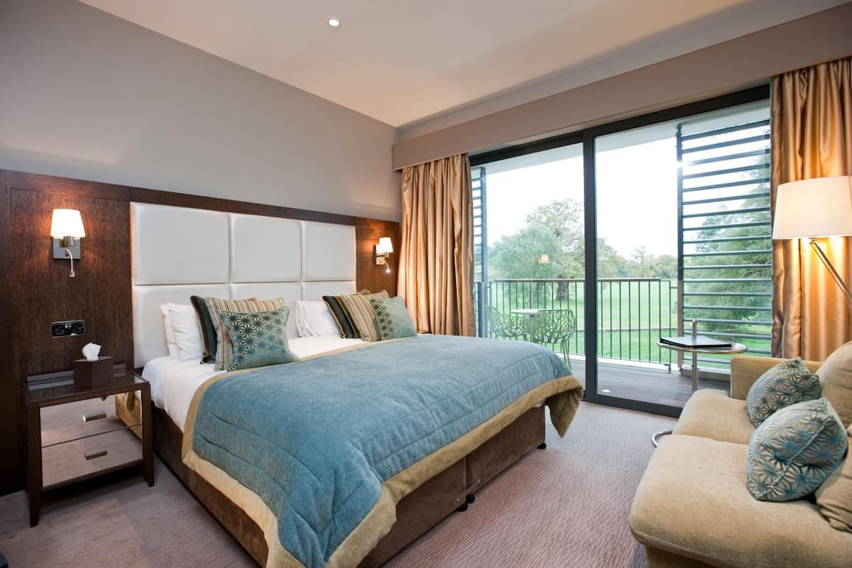 A hotel room with a large bed, a nightstand, a sofa, and a balcony with a table and chairs.