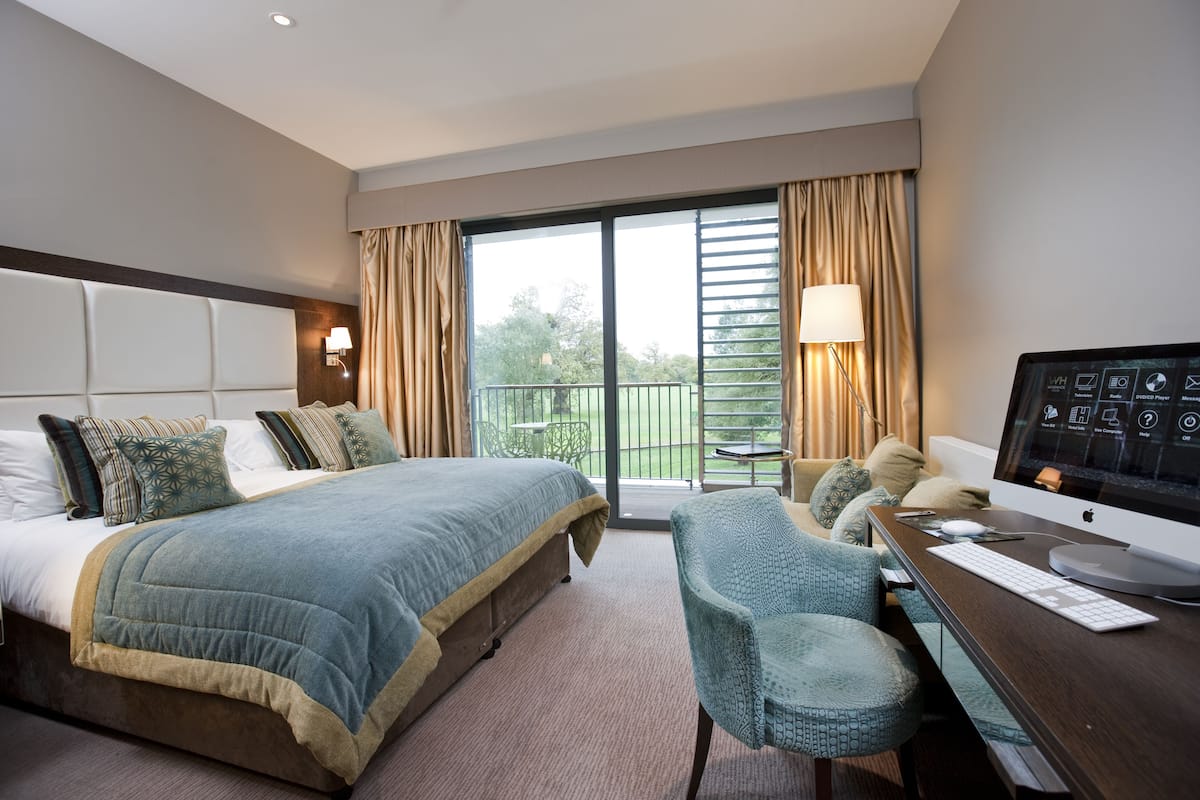 A hotel room with a bed, a desk with a computer, a chair, and a balcony with a view of greenery.