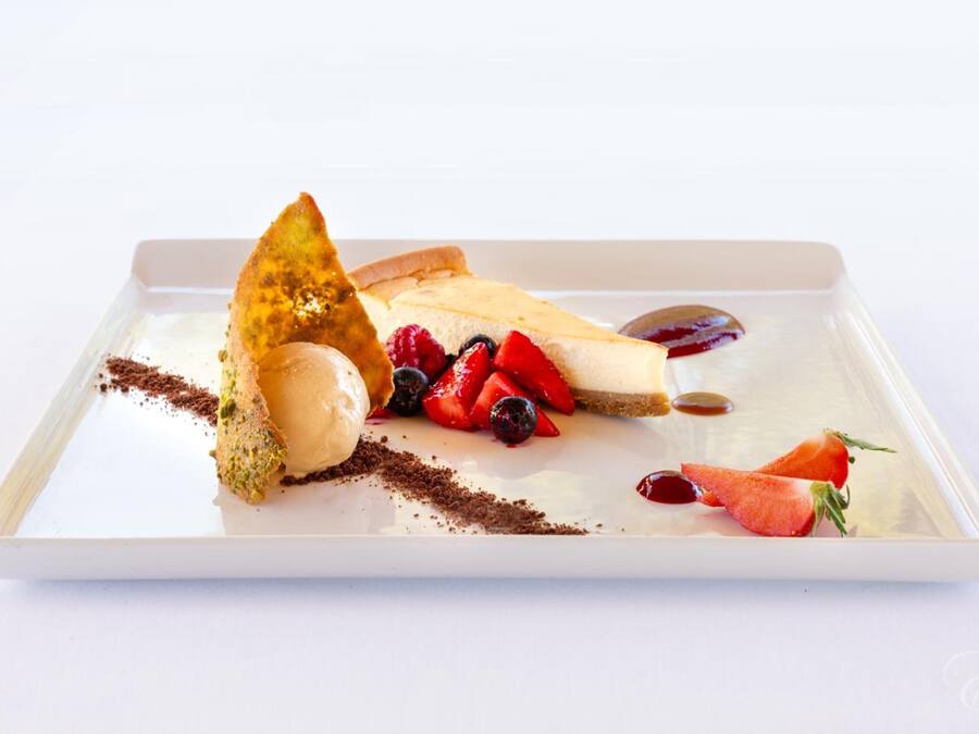 Un plato de postre con una porción de pastel de queso, fresas, arándanos y un trozo de chocolate sobre una bandeja blanca.