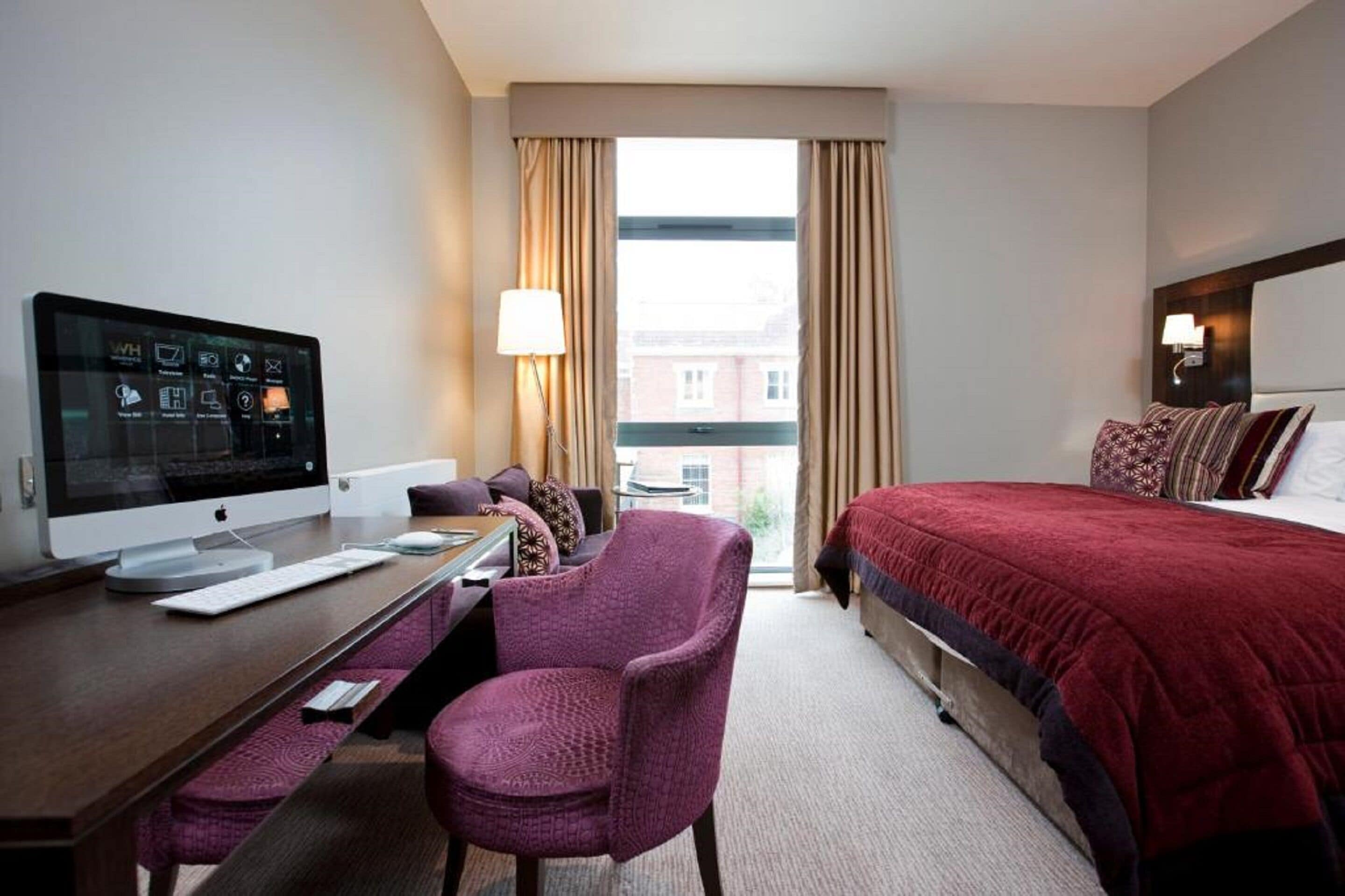 A hotel room with a bed, a desk with a computer, a chair, and a window with curtains.