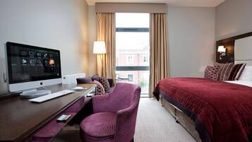 A hotel room with a bed, a desk with a computer, a chair, and a window with curtains.