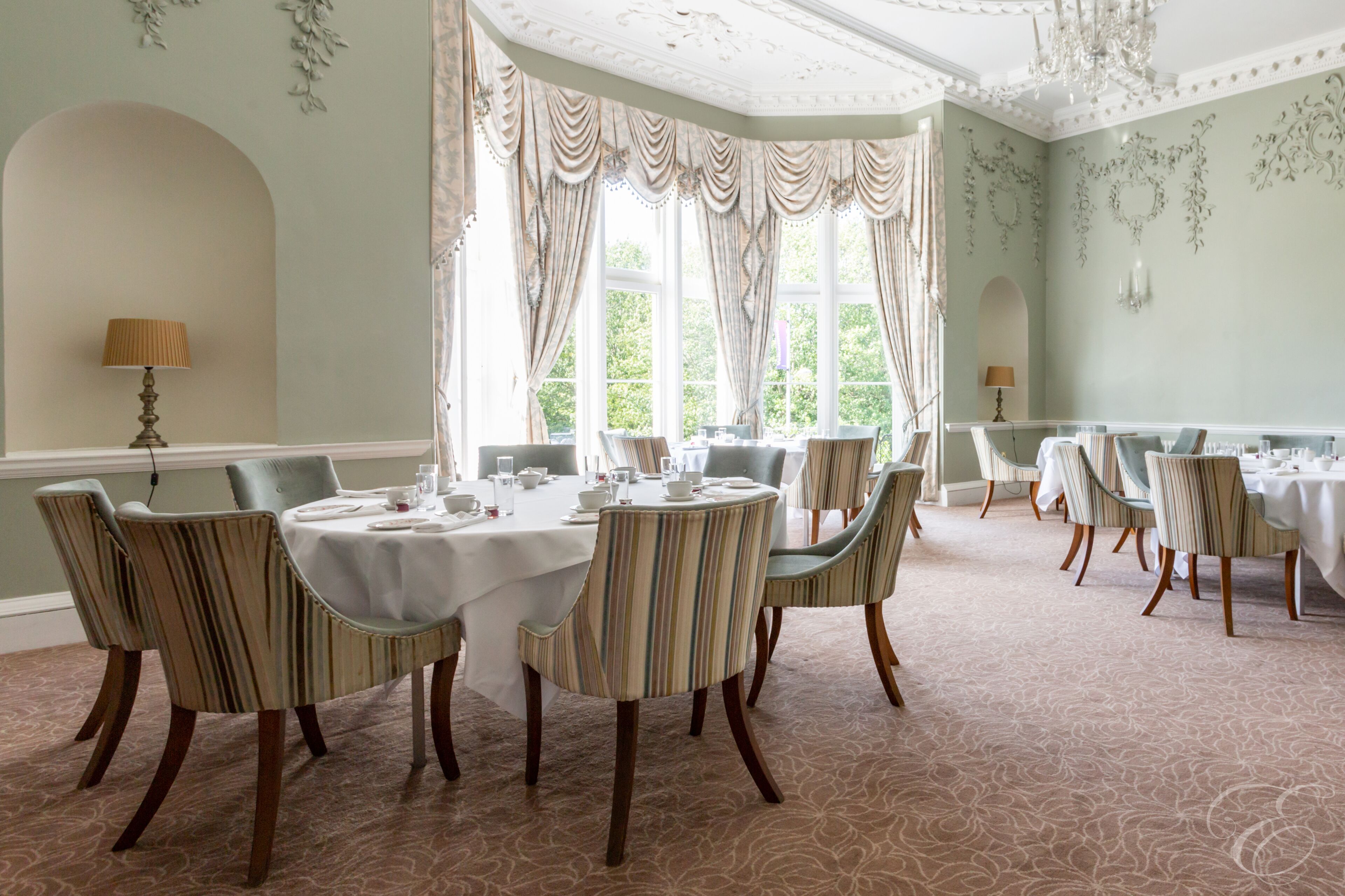 A dining room with round tables and striped chairs, large windows with curtains, and a patterned carpet.