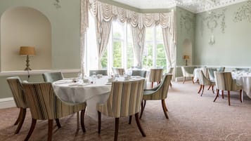 A dining room with round tables and striped chairs, large windows with curtains, and a patterned carpet.