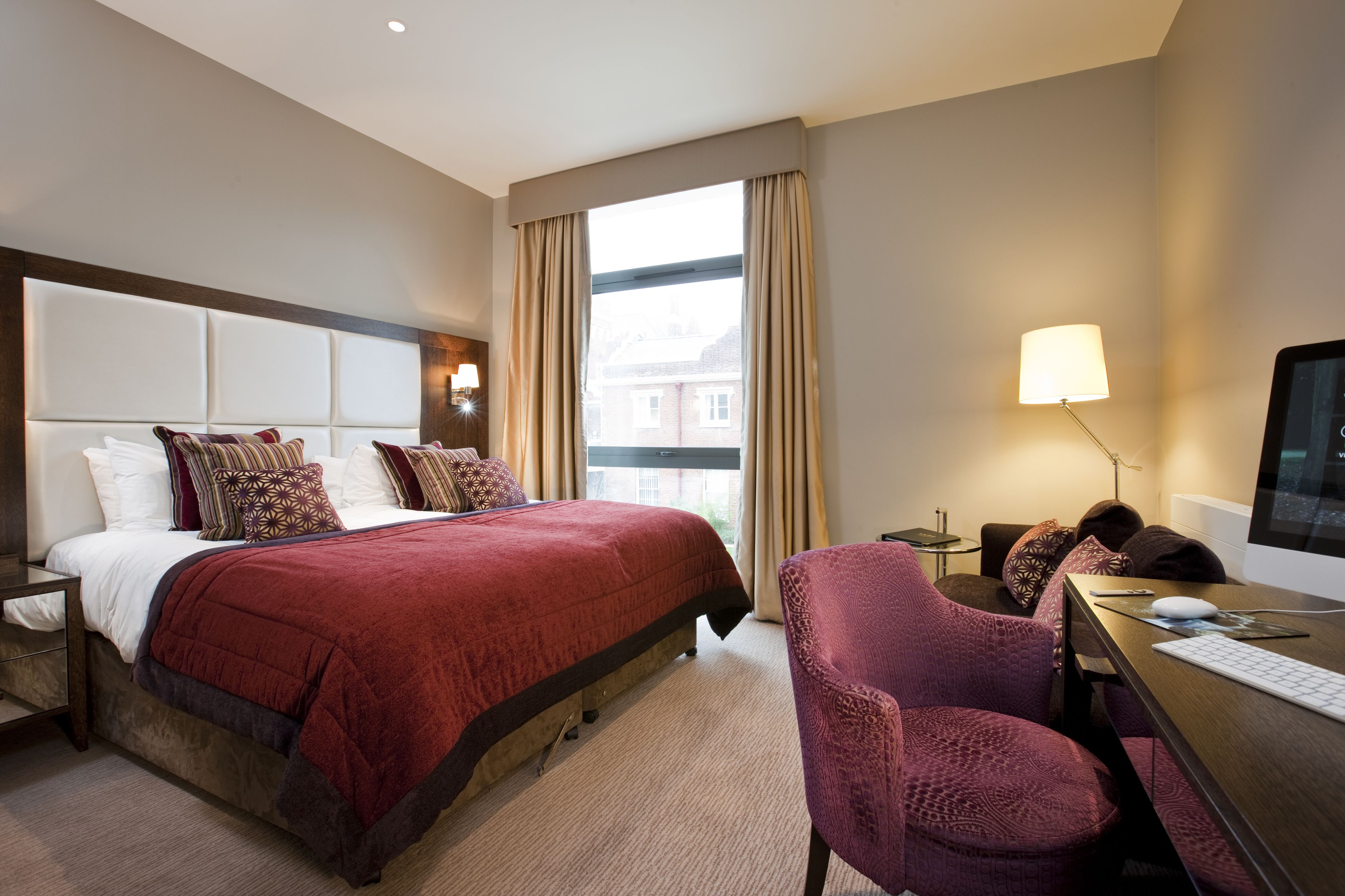 A hotel room with a large bed, a desk with a computer, a chair, and a window with curtains.