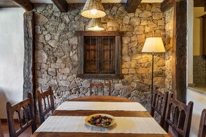 A dining area with a stone wall, wooden table, chairs, a lamp, and a window with shutters.