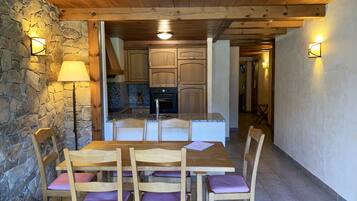 A wooden dining table with six chairs, a stone wall, and a kitchen in the background.
