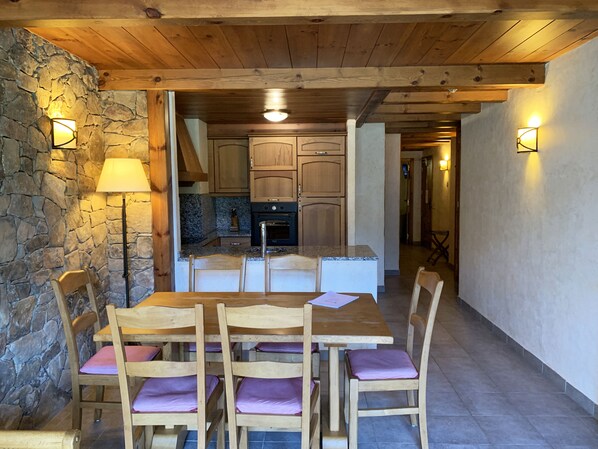 A wooden dining table with six chairs, a stone wall, and a kitchen in the background. - Apartaments Turístics Els Llacs (Bordes d'Envalira)