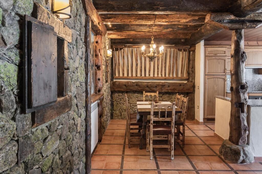 A rustic stone-walled dining area with a wooden table and chairs, a chandelier, and a stone fireplace.