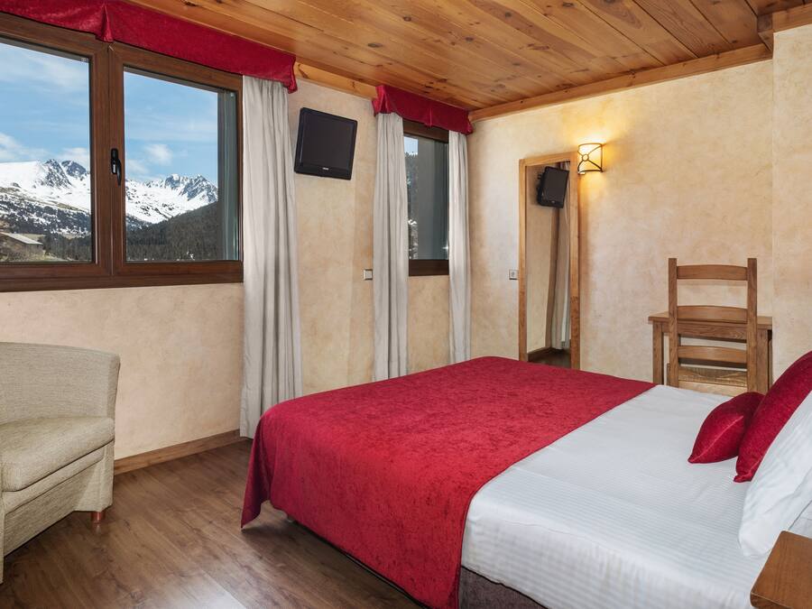 A bedroom with a bed, a chair, a window with curtains, a wall-mounted TV, and a nightstand.