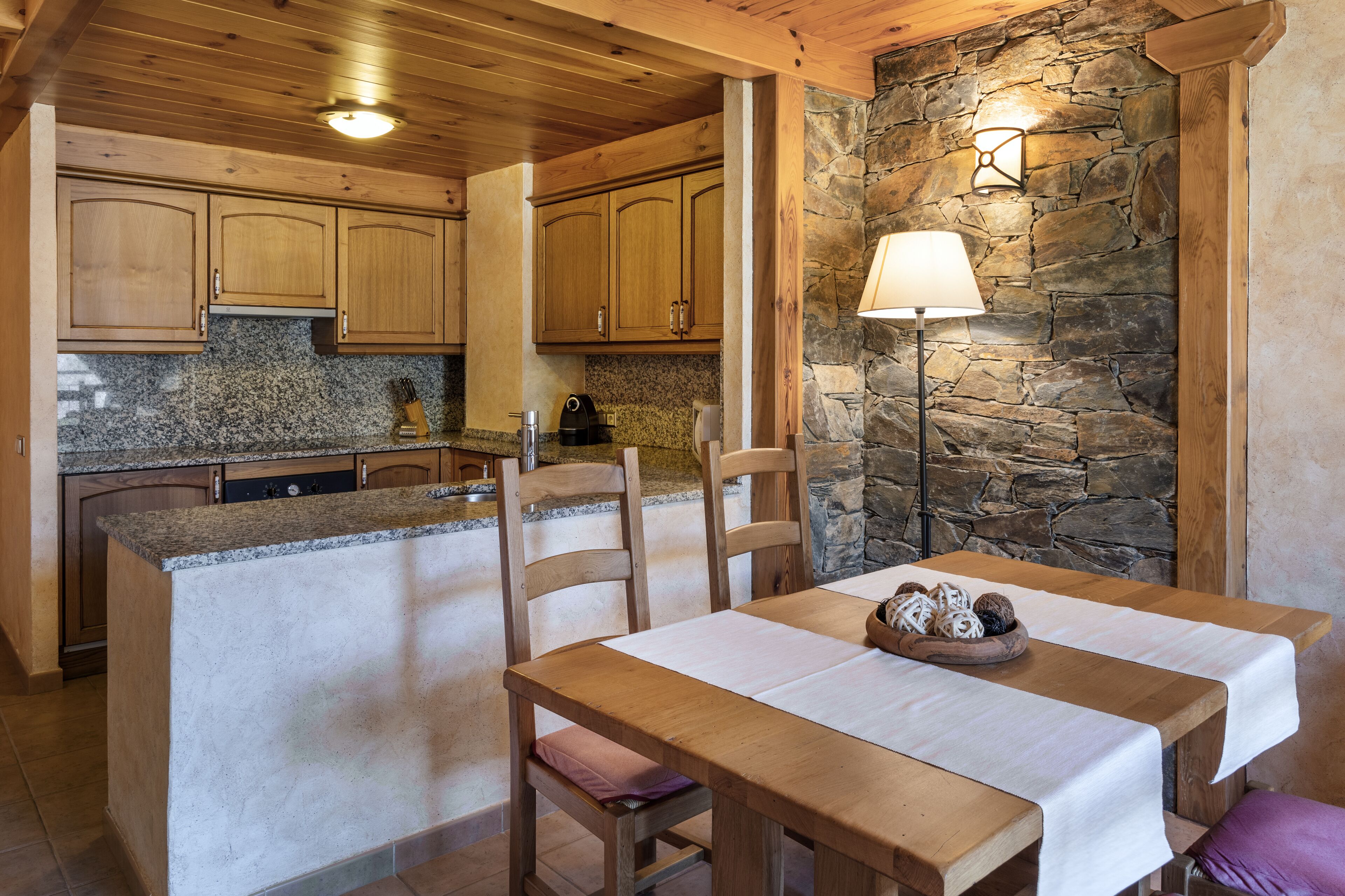 Une cuisine dotée de meubles en bois, de plans de travail en granit, d’un mur en pierre et d’un coin repas avec une table mise pour deux personnes.