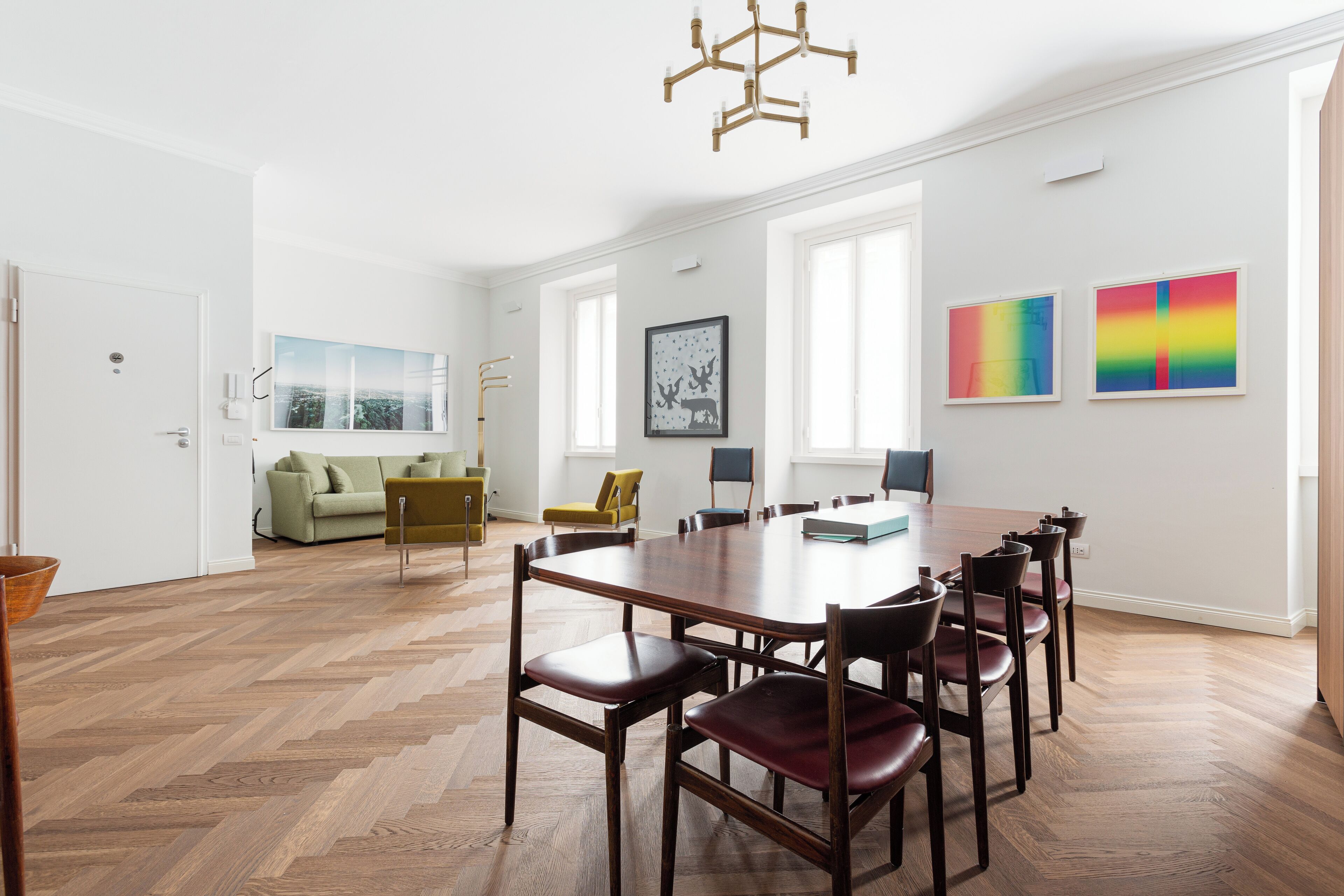A spacious room with a large wooden dining table surrounded by chairs, a sofa, and a yellow armchair. There are large windows providing natural light, and artwork is displayed on the walls.