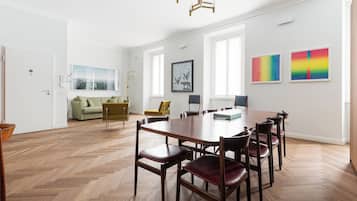 A spacious room with a large wooden dining table surrounded by chairs, a sofa, and a yellow armchair. There are large windows providing natural light, and artwork is displayed on the walls.