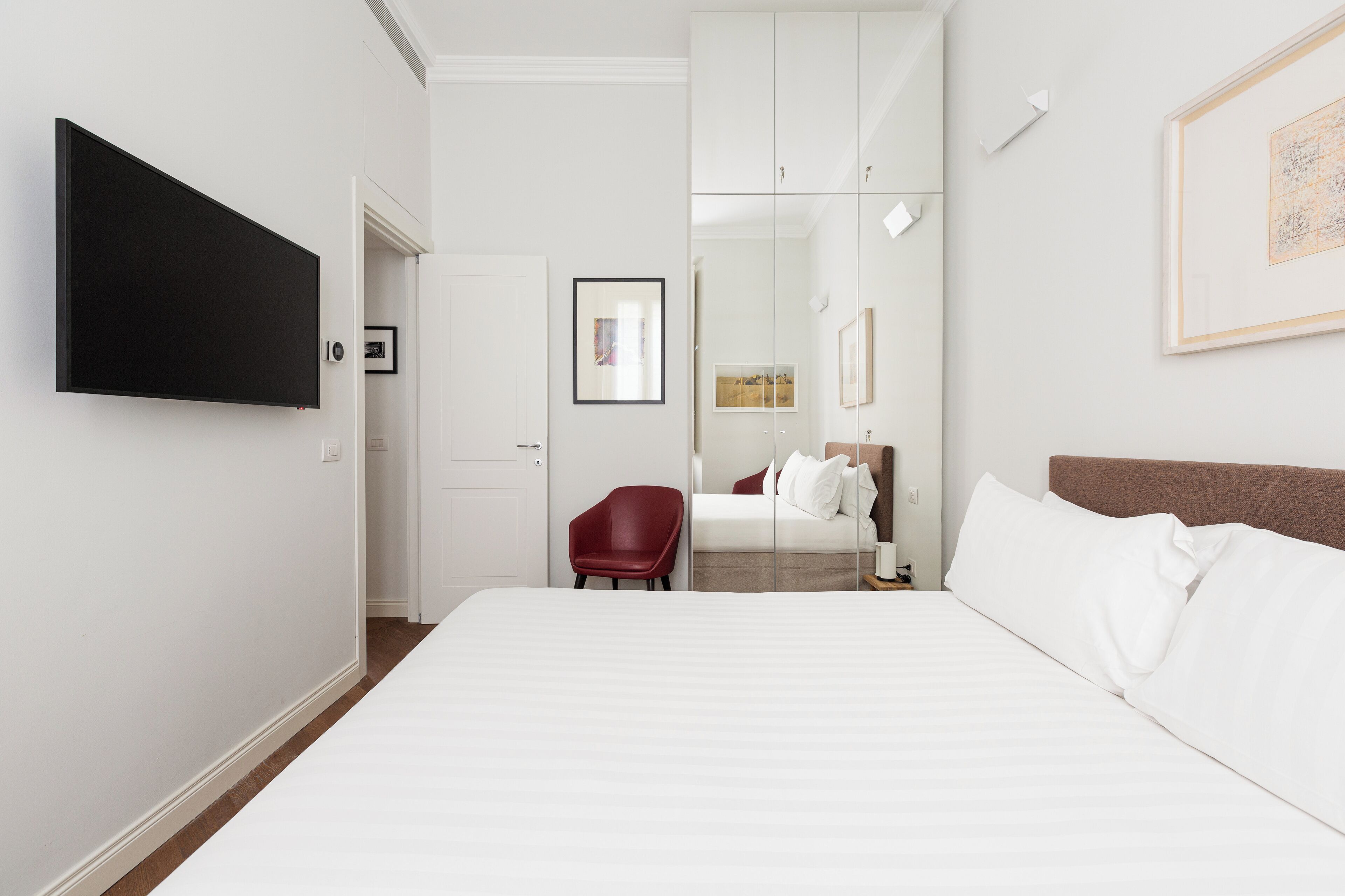 A hotel room with a large bed, a red chair, a flat-screen TV mounted on the wall, and a framed picture above the bed.