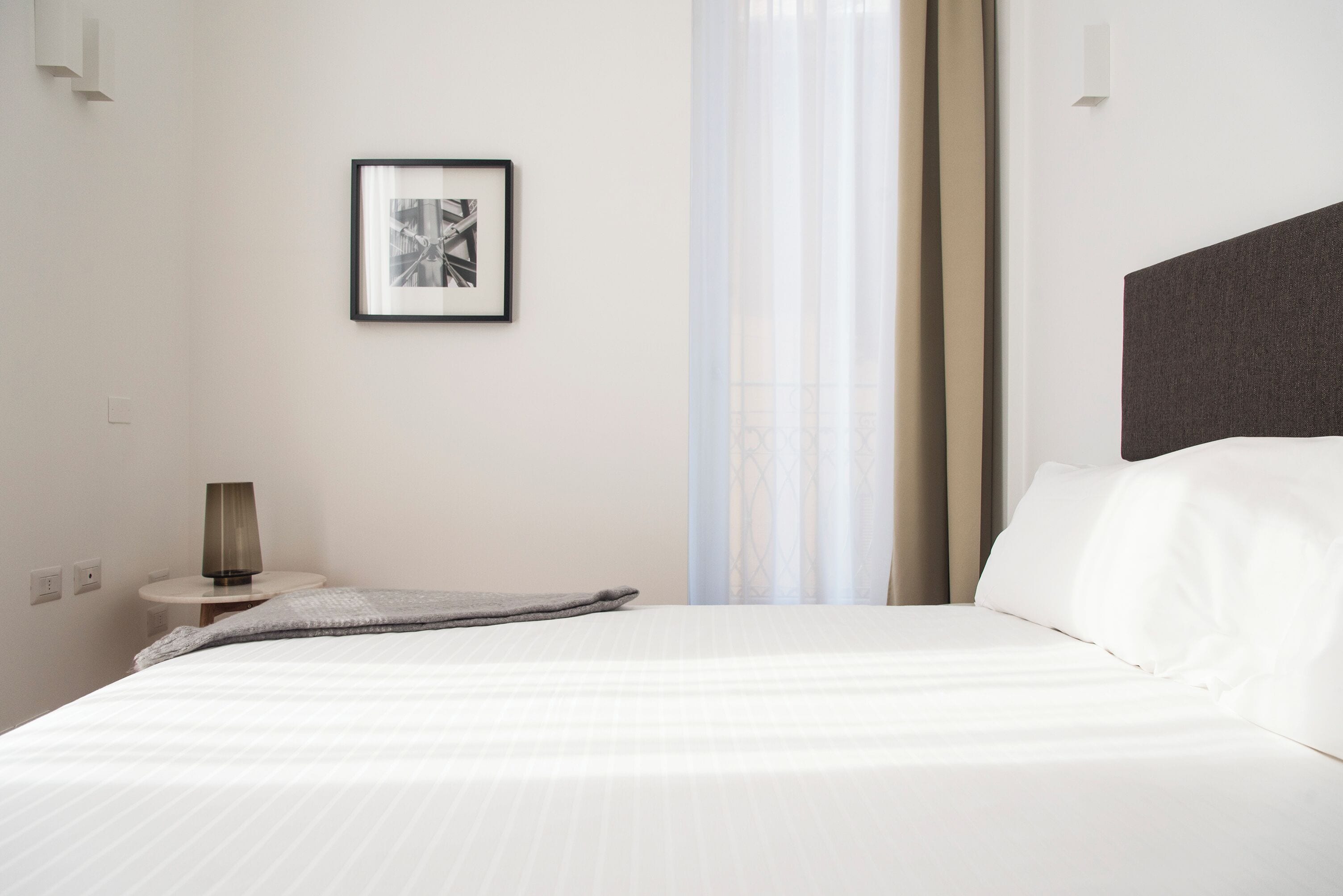 Una cama bien hecha con sábanas blancas, una mesita de noche con lámpara, un cuadro en la pared y una cortina translúcida entreabierta.