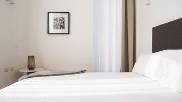 Una cama bien hecha con sábanas blancas, una mesita de noche con lámpara, un cuadro en la pared y una cortina translúcida entreabierta.