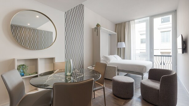 A modern hotel room with a round glass table, two chairs, a bed, a sofa, and a large window. - Brera Apartments in San Babila (Milan)