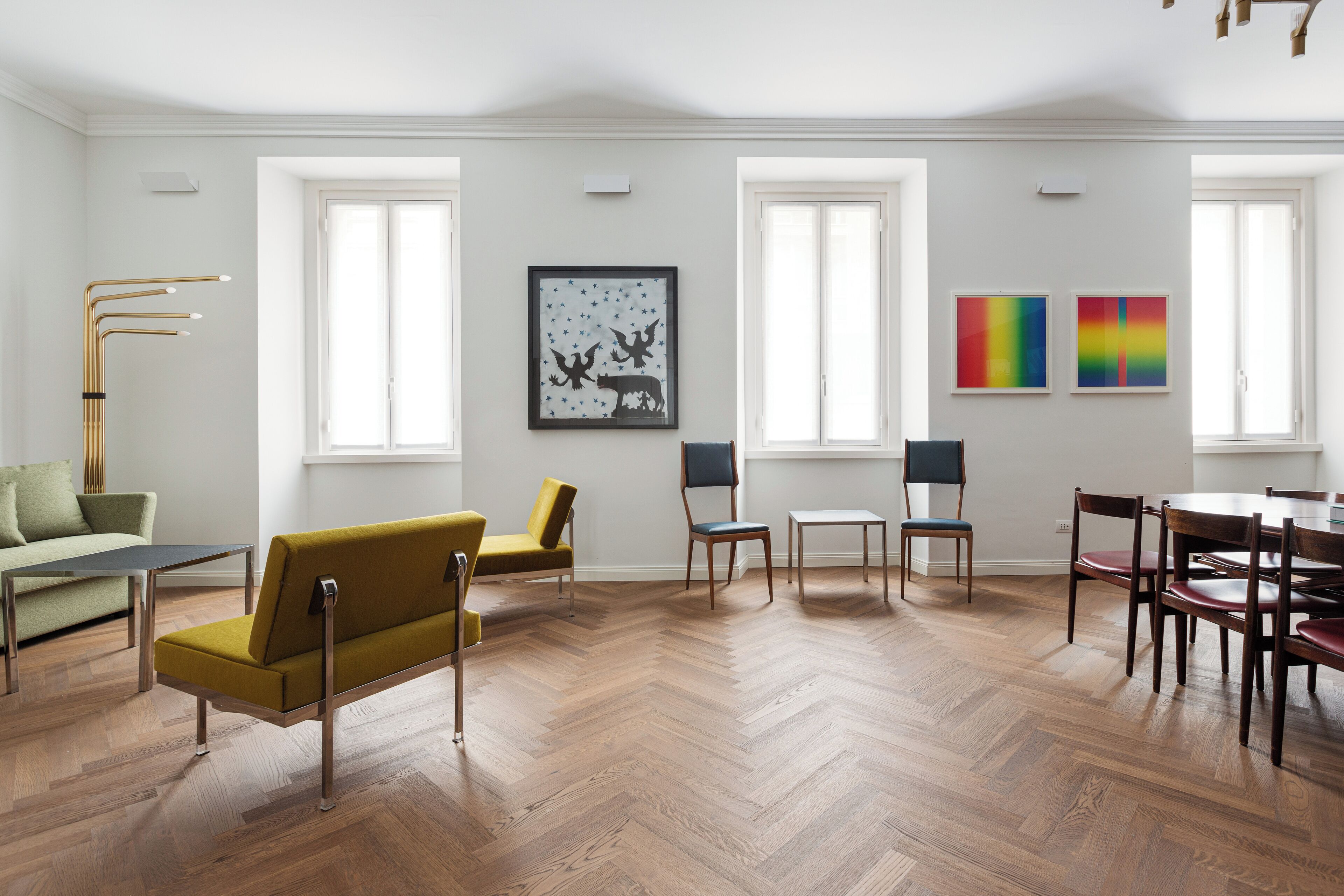 Une pièce spacieuse dotée d’un parquet en bois à motifs chevrons, d’un canapé vert, d’un fauteuil jaune, d’une table à manger avec des chaises et de deux fenêtres à cadres blancs.