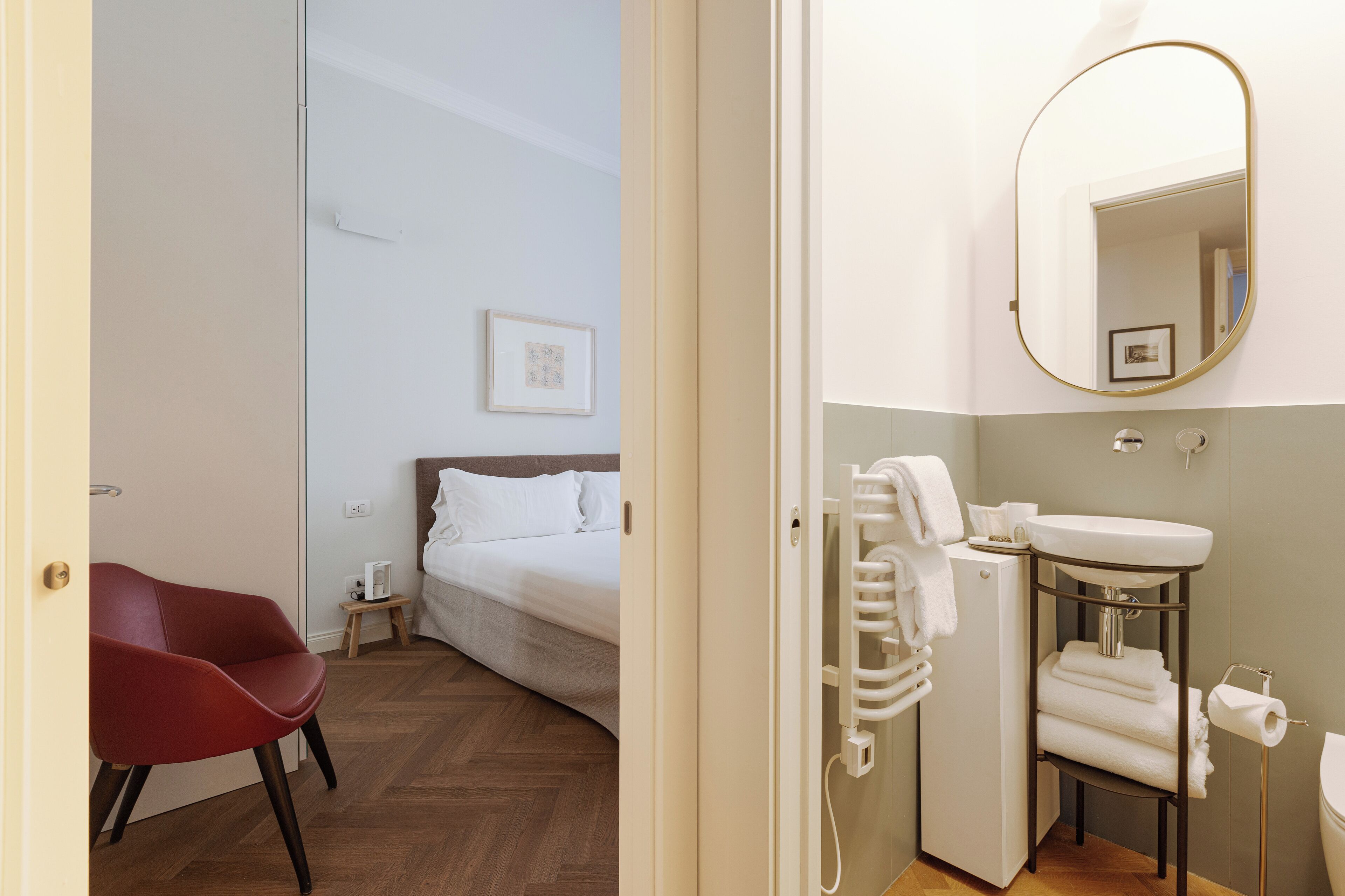 Una habitación de hotel con cama, una silla roja, una mesita de noche pequeña, un lavamanos blanco y un espejo.