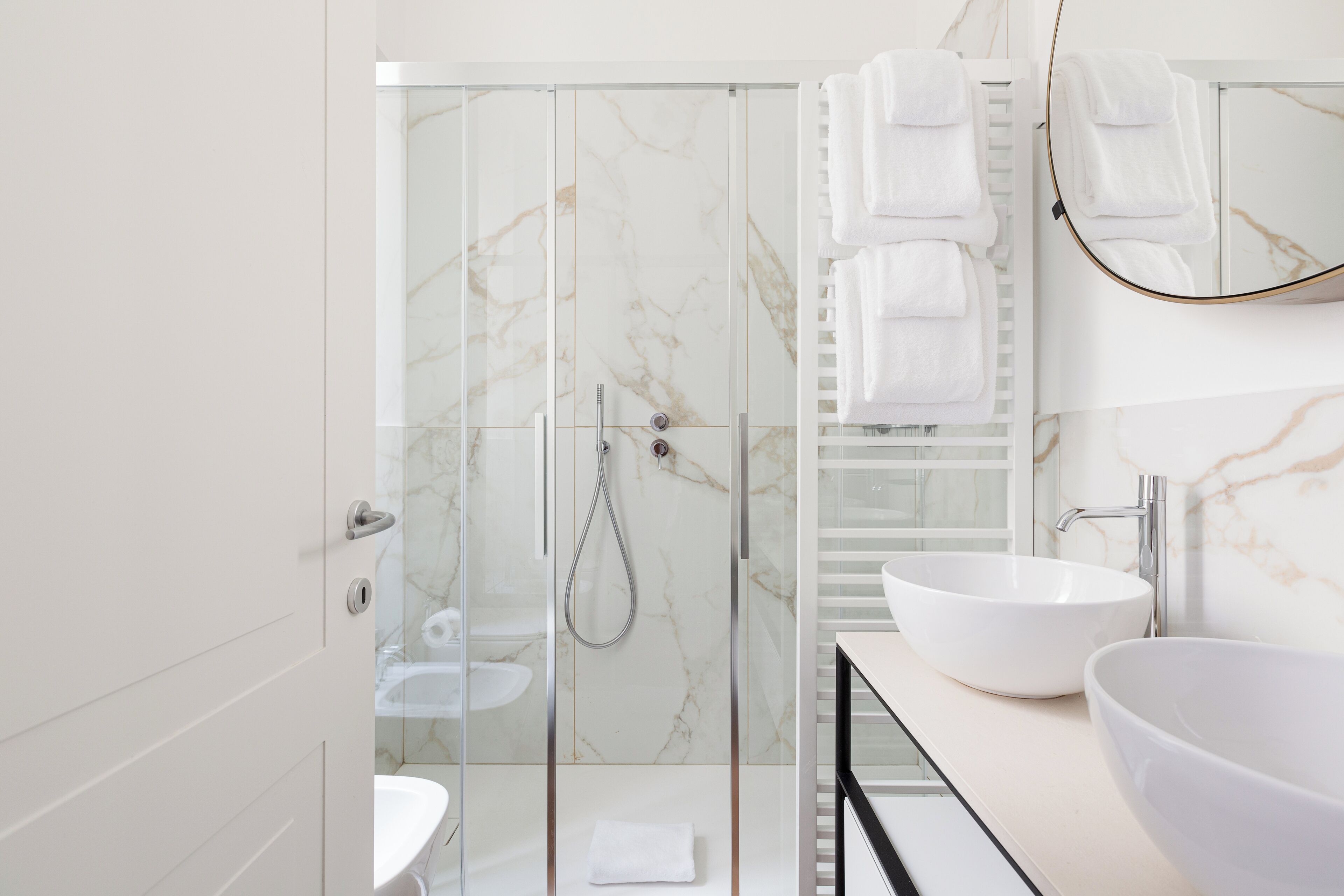 Un baño moderno con ducha envuelta en vidrio, dos lavamanos blancos y un salpicadero de mármol.