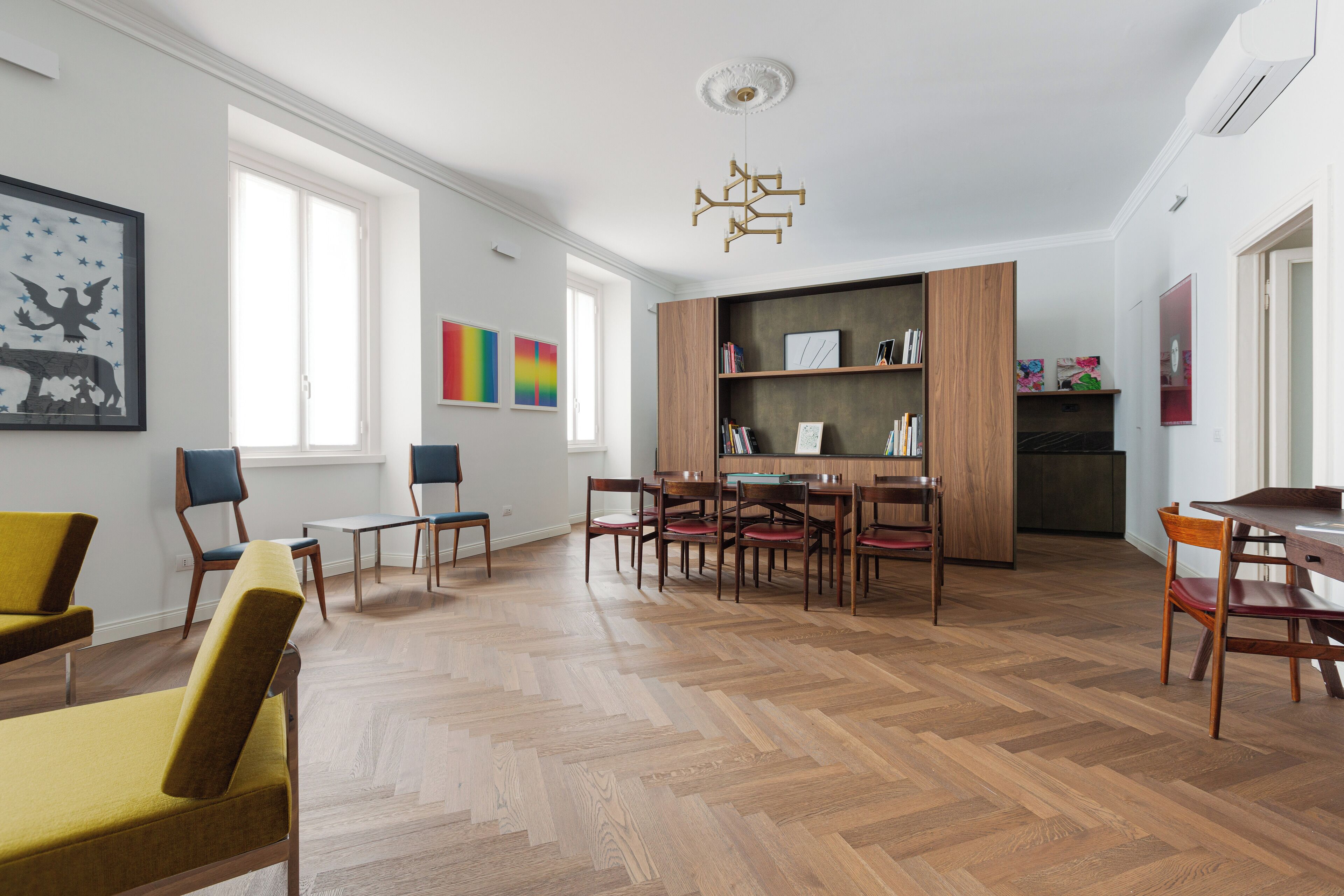 A spacious living area with a herringbone wood floor, a dining table with chairs, a built-in bookshelf, and a ceiling light fixture.