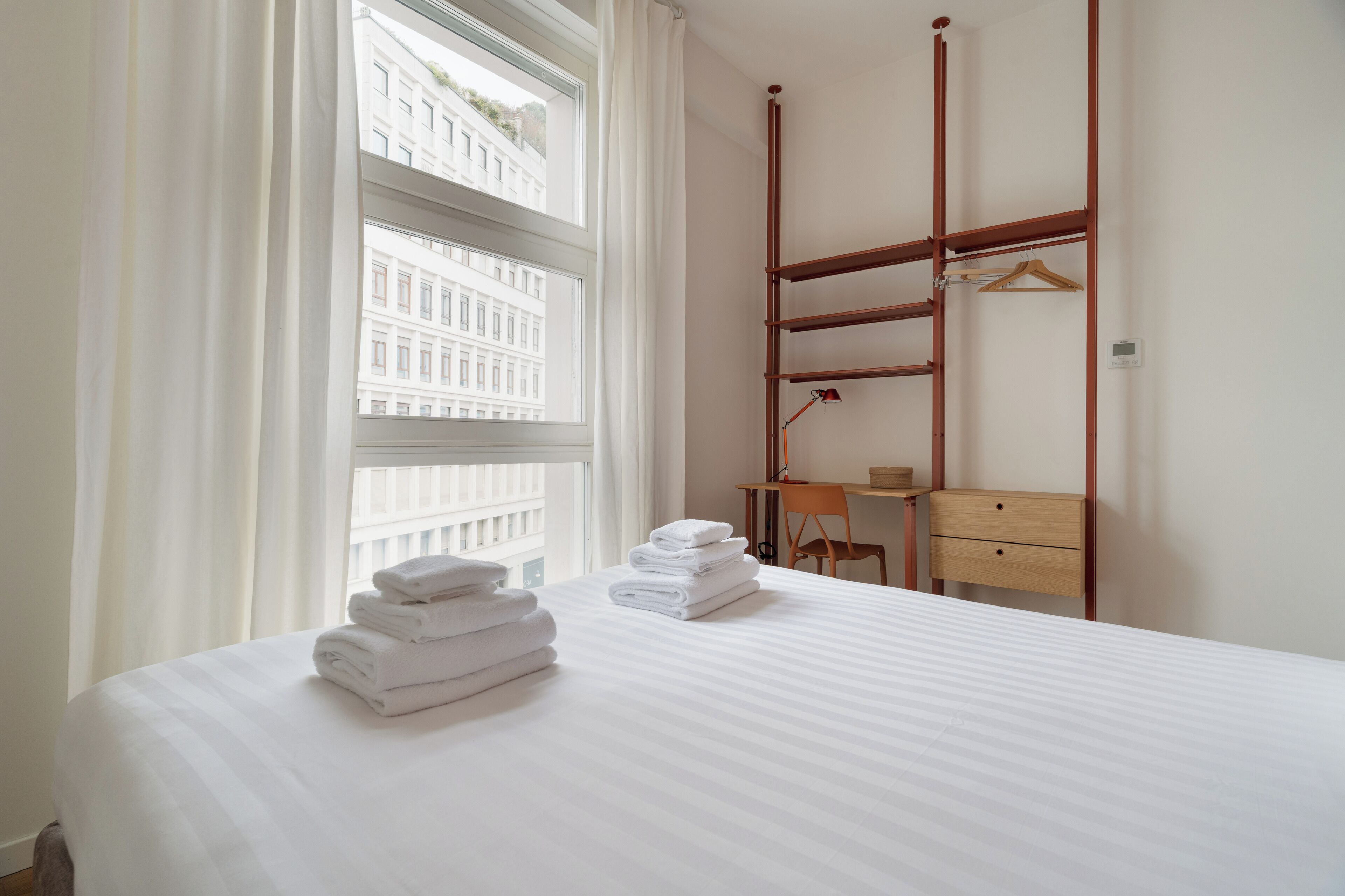 A hotel room with a bed, a desk, a chair, a wooden wardrobe, and a window with curtains.