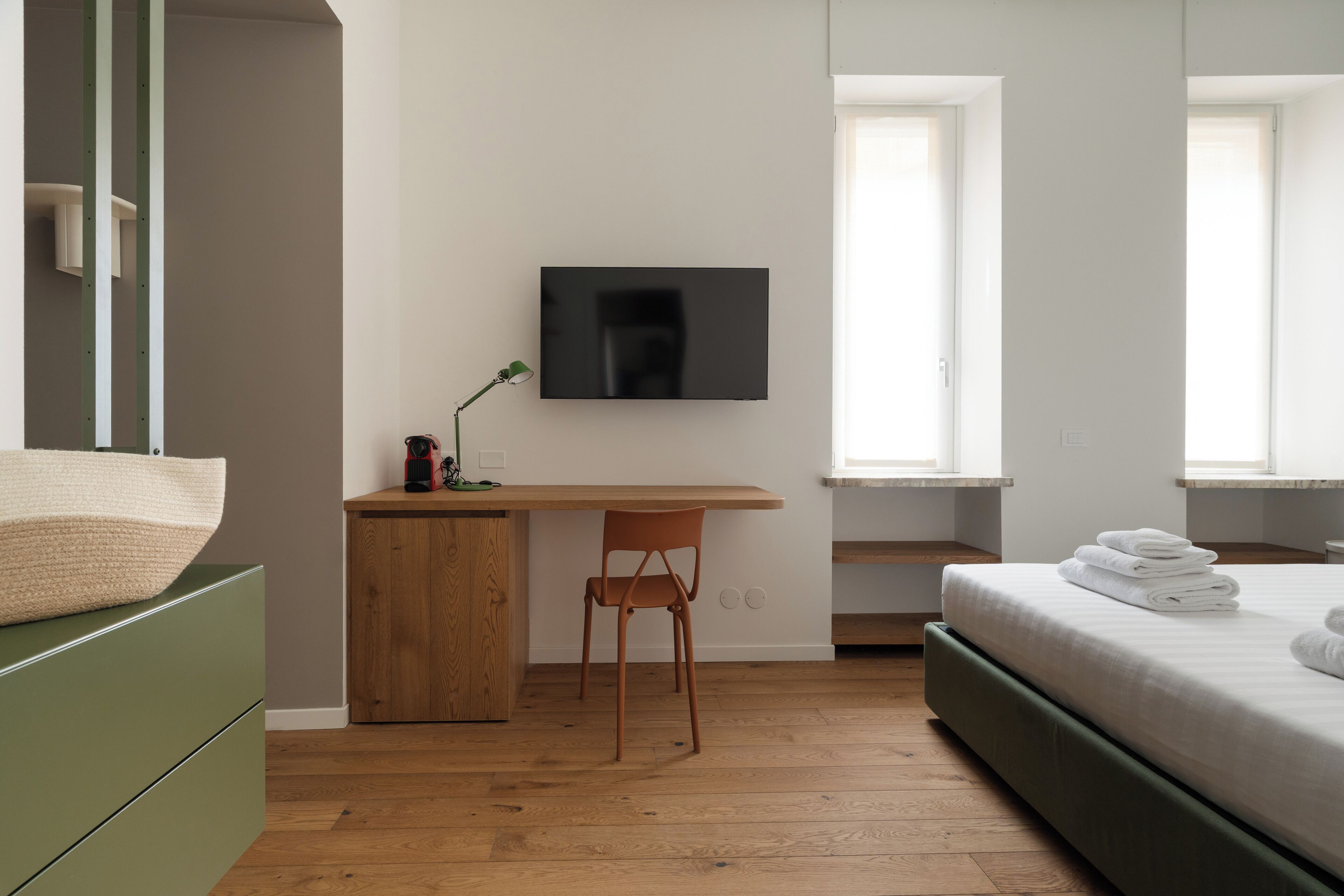 A bedroom with a bed, a desk with a chair, a TV mounted on the wall, and a window.