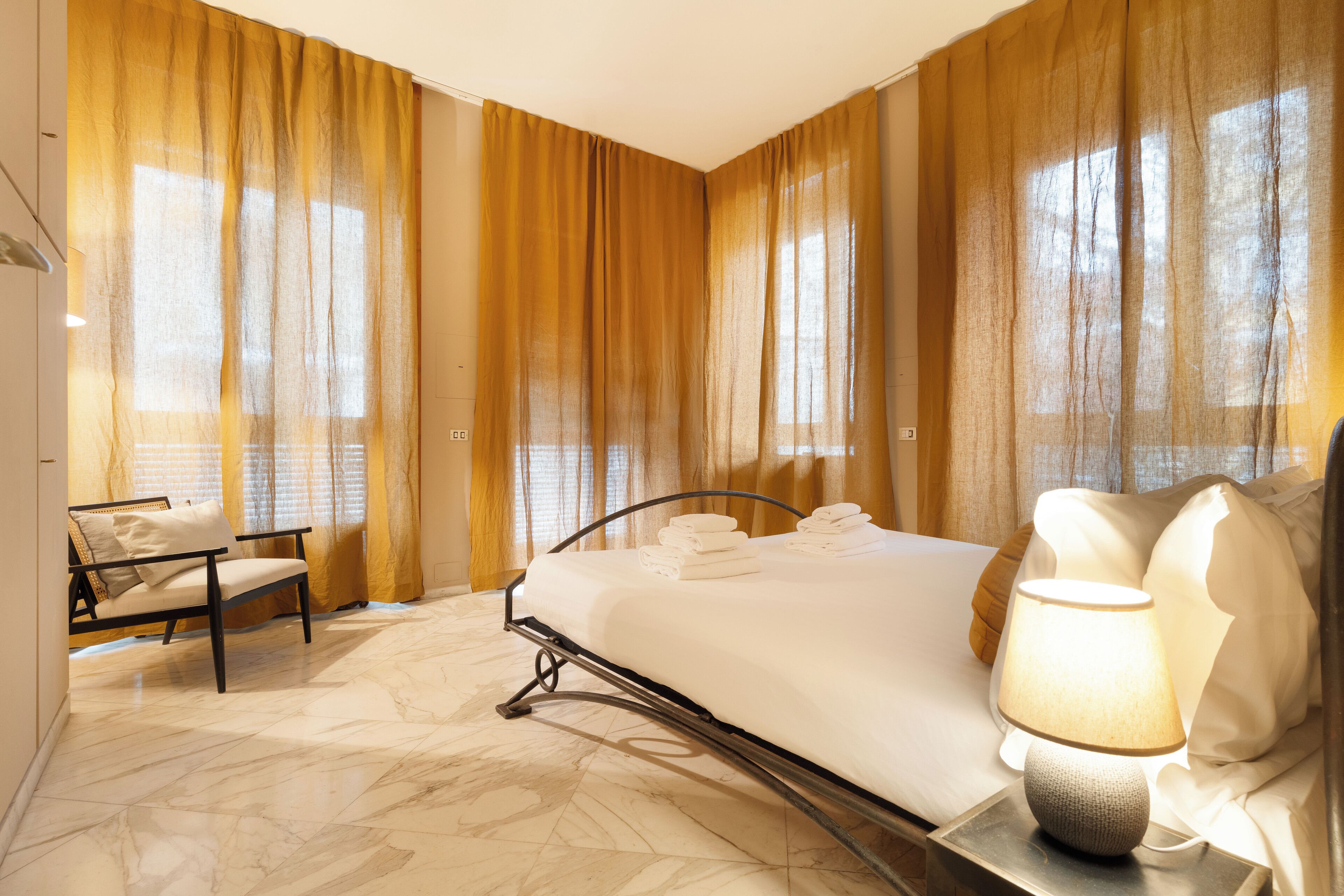 A hotel room with a bed, a chair, a lamp, and large windows with curtains.
