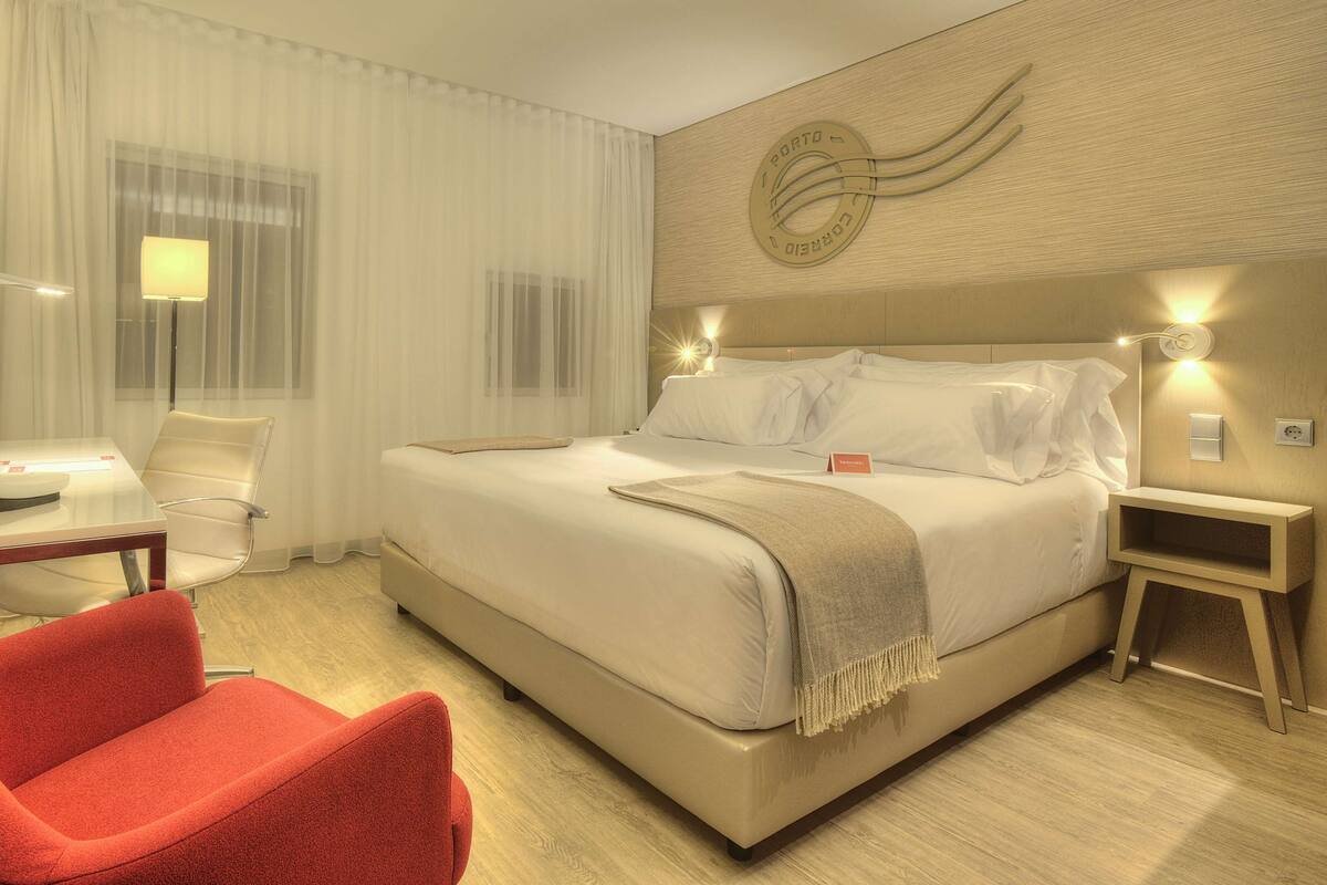 A hotel room with a large bed, a red armchair, a desk with a lamp, and a wall-mounted TV.