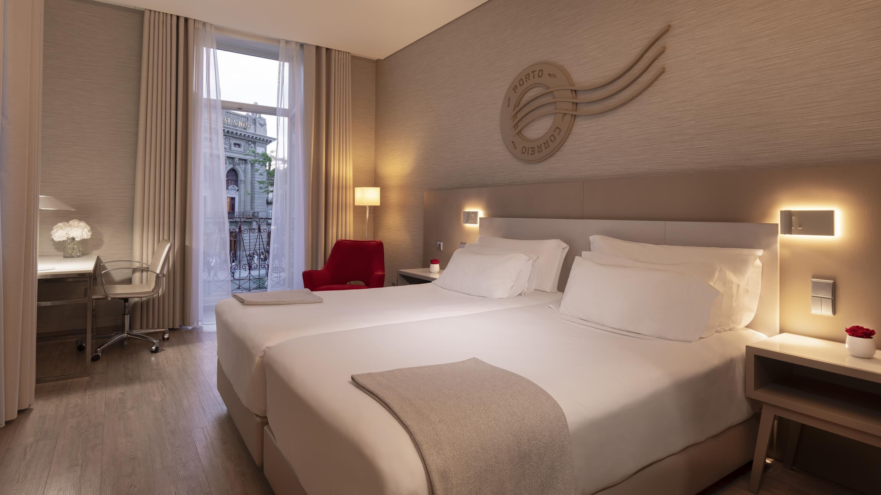 Een hotelkamer met een groot bed, een bureau met een stoel, een rode stoel en uitzicht op een gebouw door het raam.