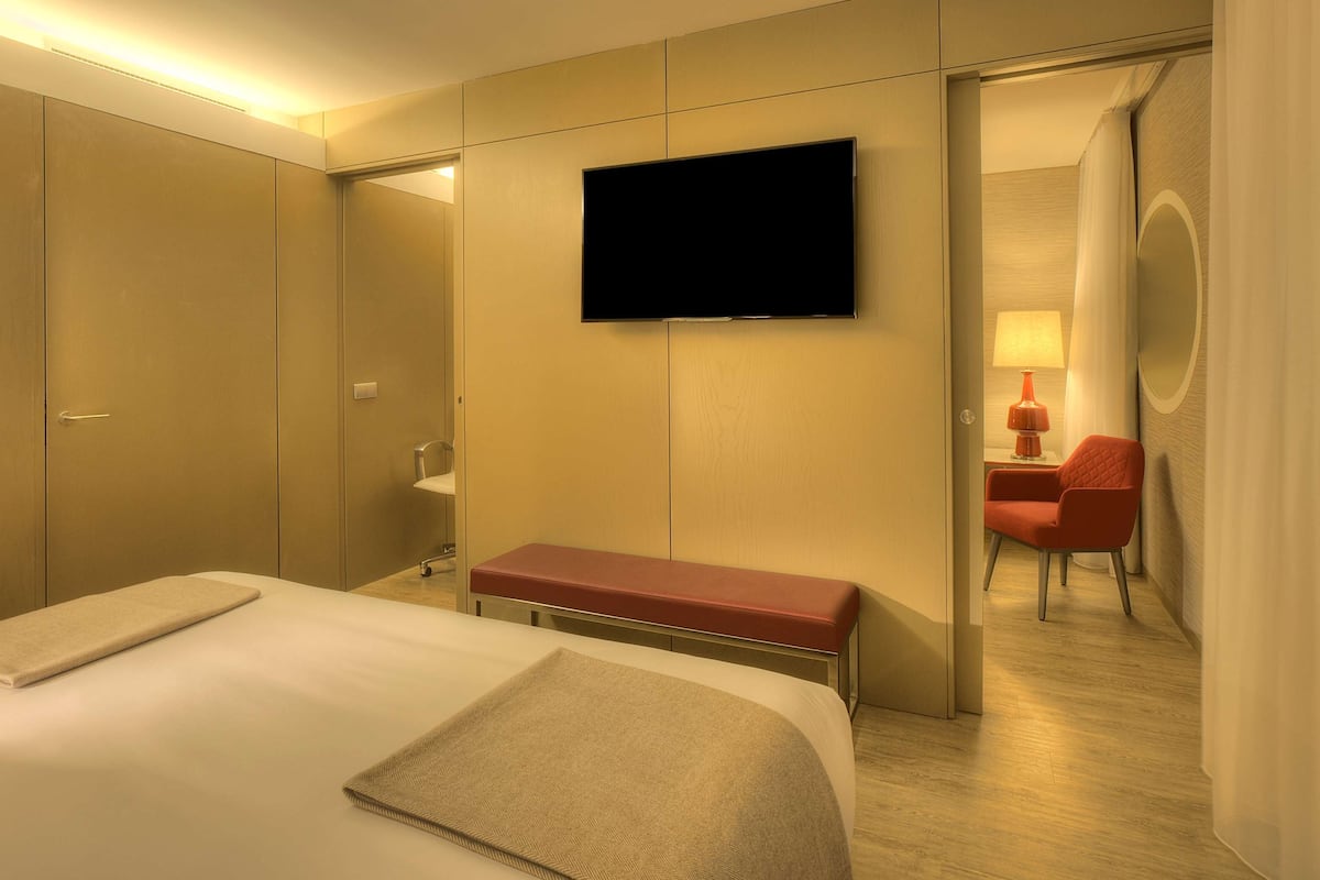 A hotel room with a bed, a bench, a TV, a desk with a chair, and a bathroom.
