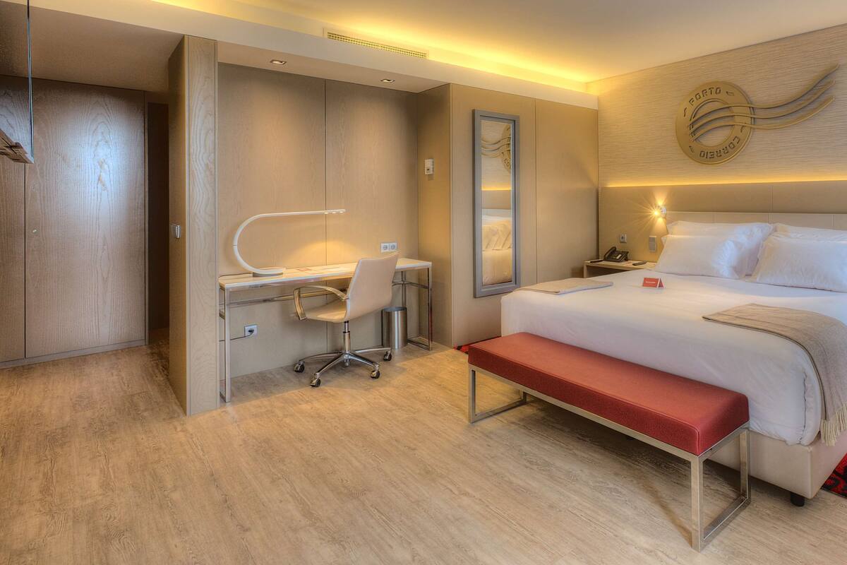 A modern hotel room with a bed, desk, chair, and a red bench.