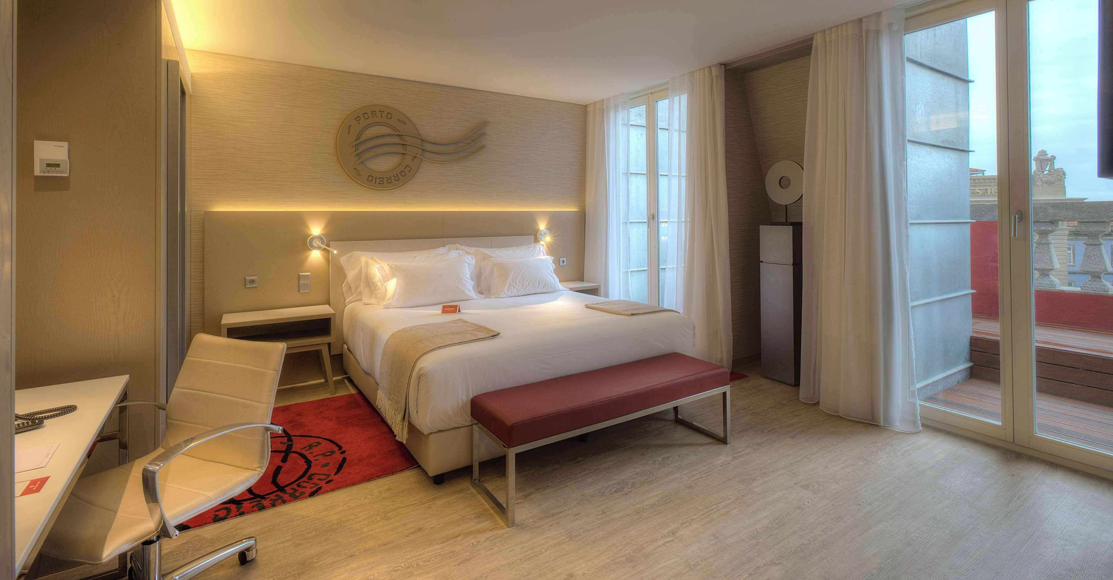 A hotel room with a bed, bedside tables, a desk, a chair, a bench, and a large window with curtains.