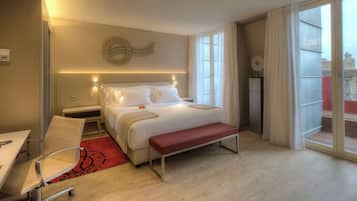 A hotel room with a bed, bedside tables, a desk, a chair, a bench, and a large window with curtains.