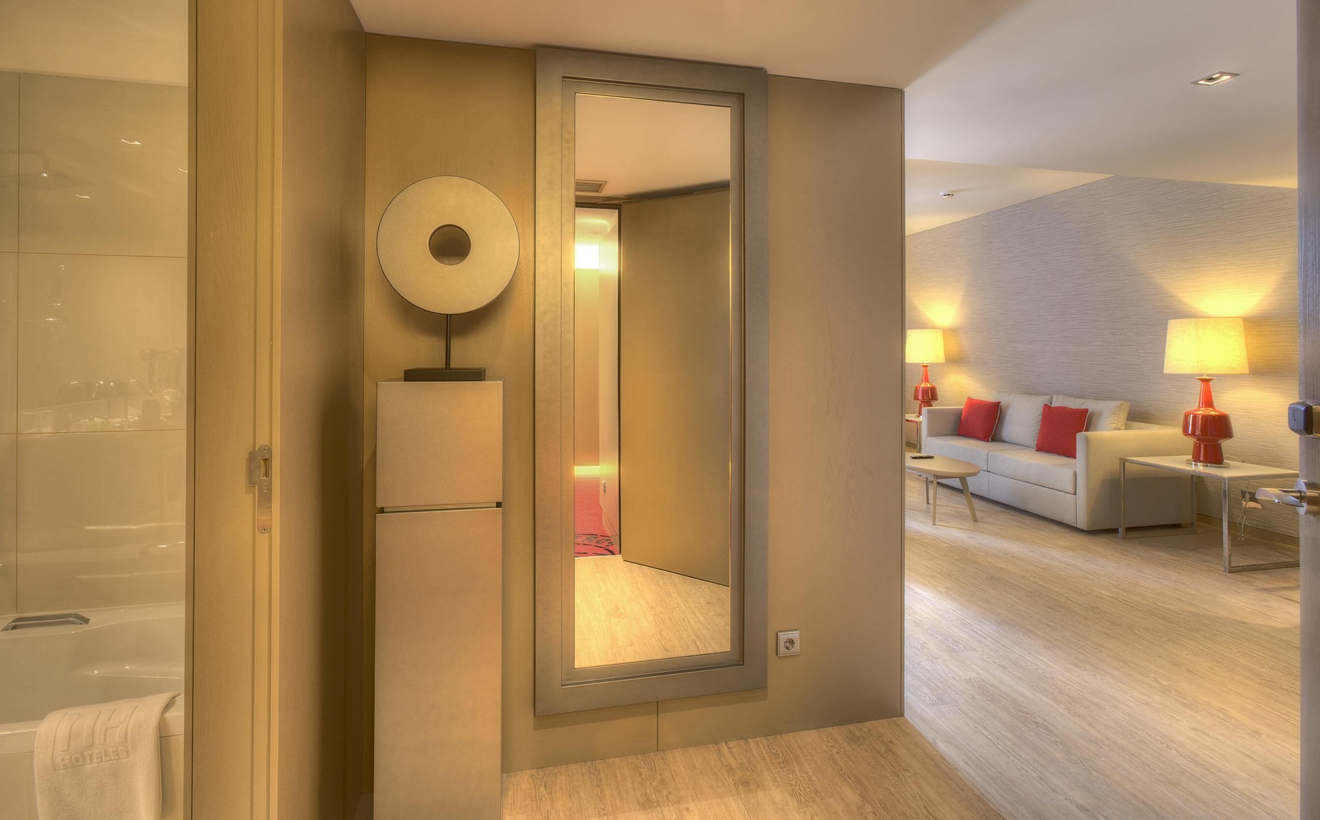 Habitación de hotel con un espejo grande, un sofá, dos mesitas de noche con lámparas y suelo de madera.