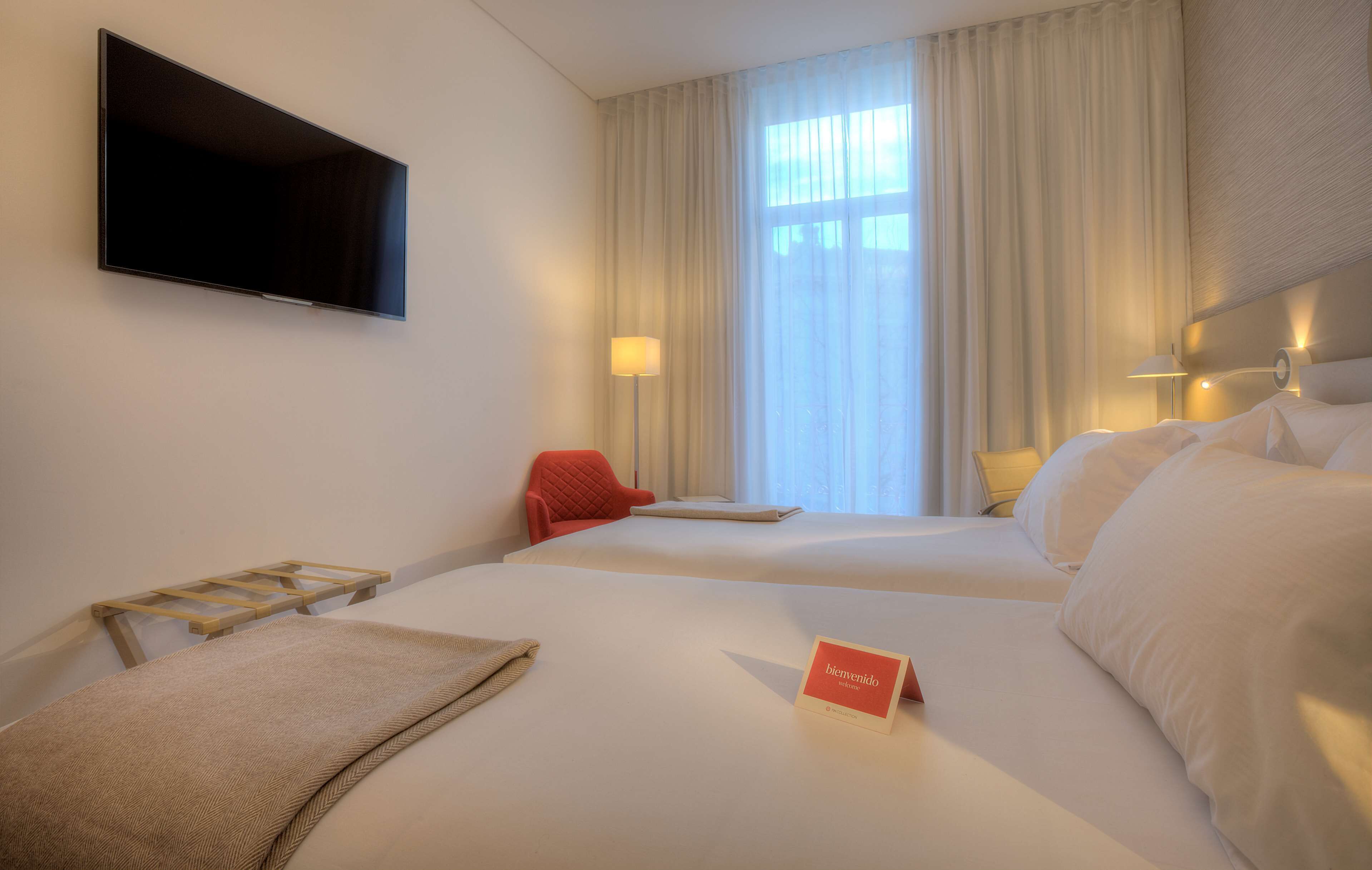 A hotel room with two beds, a flat-screen TV, a desk with a lamp, and a window with curtains.