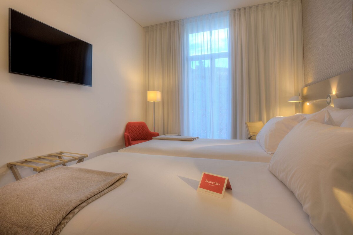 Una habitación de hotel con dos camas, un televisor de pantalla plana, un escritorio con l ámpara y un ventanal con cortinas.