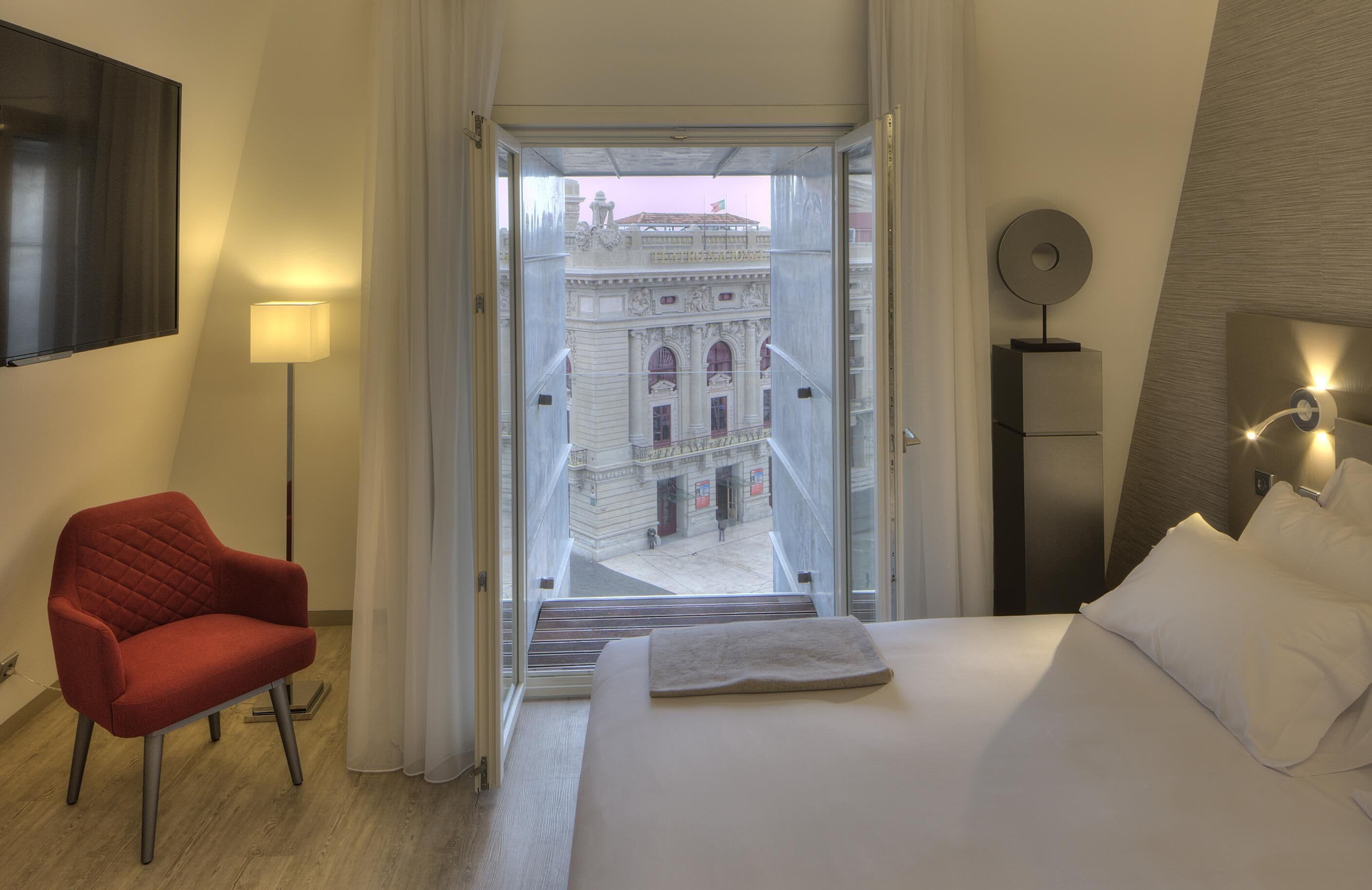 A hotel room with a bed, a red chair, a lamp, and a view of a historic building through the window.