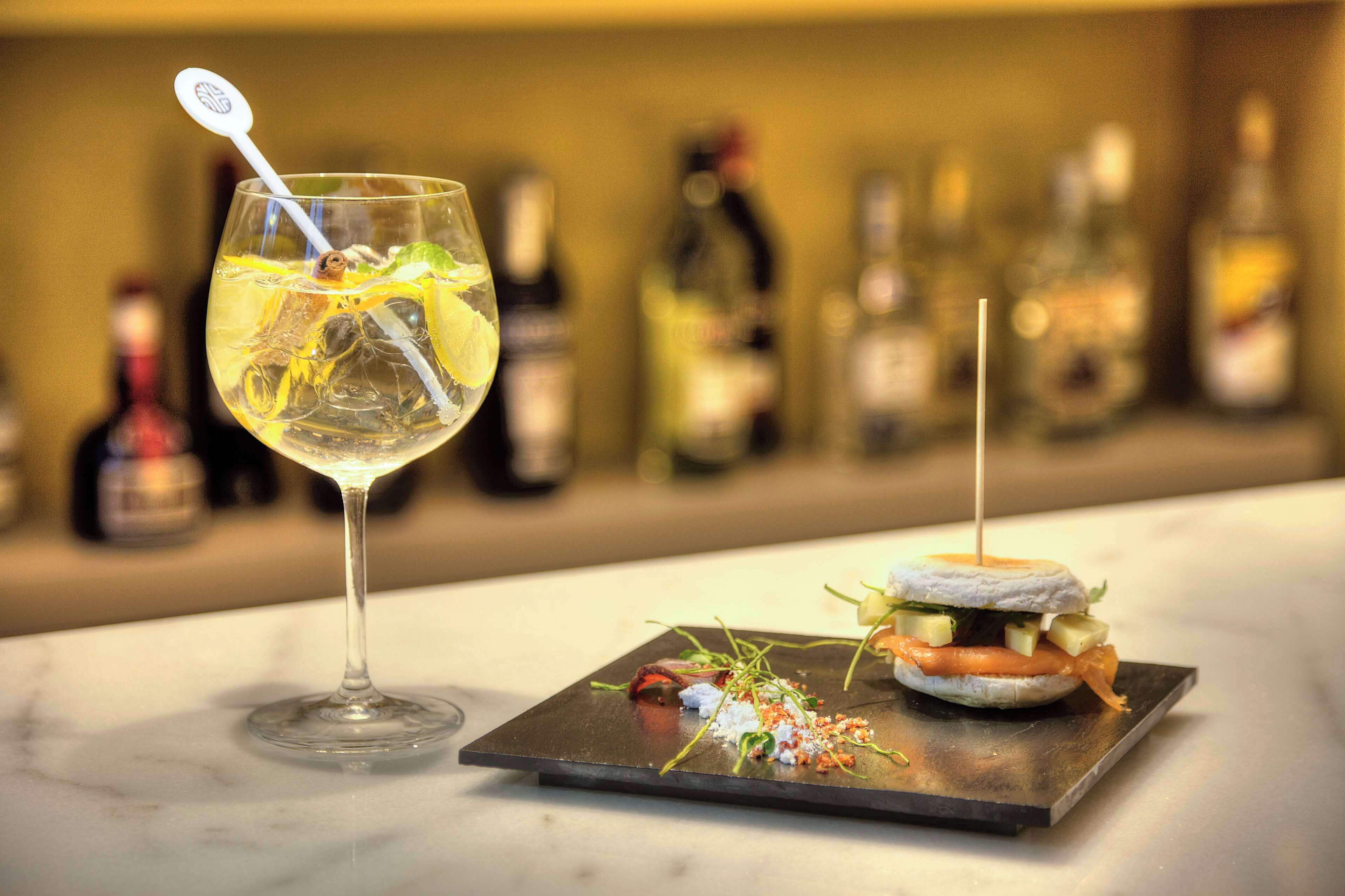 A martini glass with a straw and lemon slice, a miniature sandwich on a slate plate, and a row of blurred bottles in the background.
