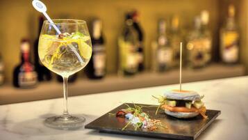 Una copa de martini con pajita y una rodaja de limón, un sándwich pequeño sobre un plato de pizarra, y una fila de botellas desenfocadas al fondo.