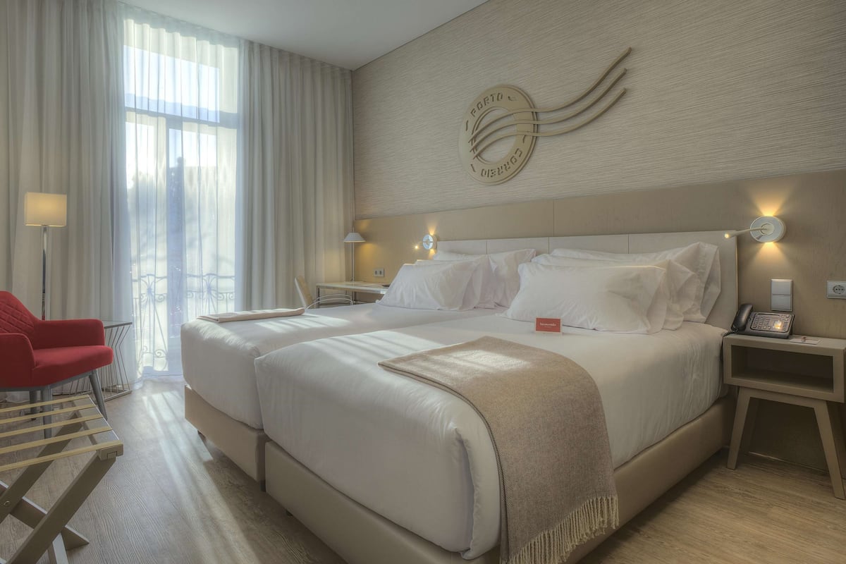 A hotel room with two beds, a red armchair, a desk, and a large window with curtains.