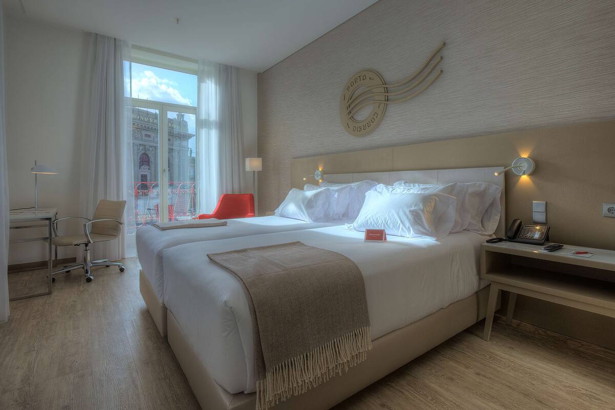 A hotel room with a large bed, a desk, a chair, and a view of a building through the window.