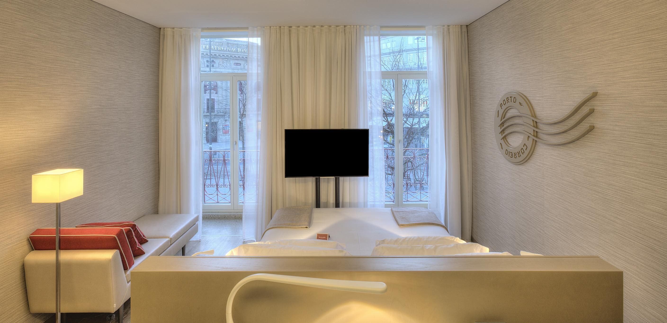 A hotel room with a bed, a TV, a bedside table, a lamp, and a window with curtains.