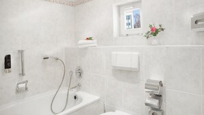 A modern bathroom with a bathtub, shower, and toilet. There is a small window with a view of trees, a towel rack, and a soap dispenser. - Kaiserhof TWO by Centro Basic (Luebeck)