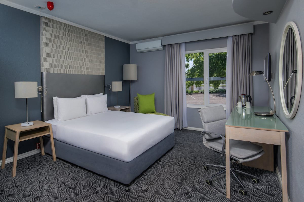 A hotel room with a large bed, a desk with a mirror, two bedside tables, a chair, and a view of the outdoors.