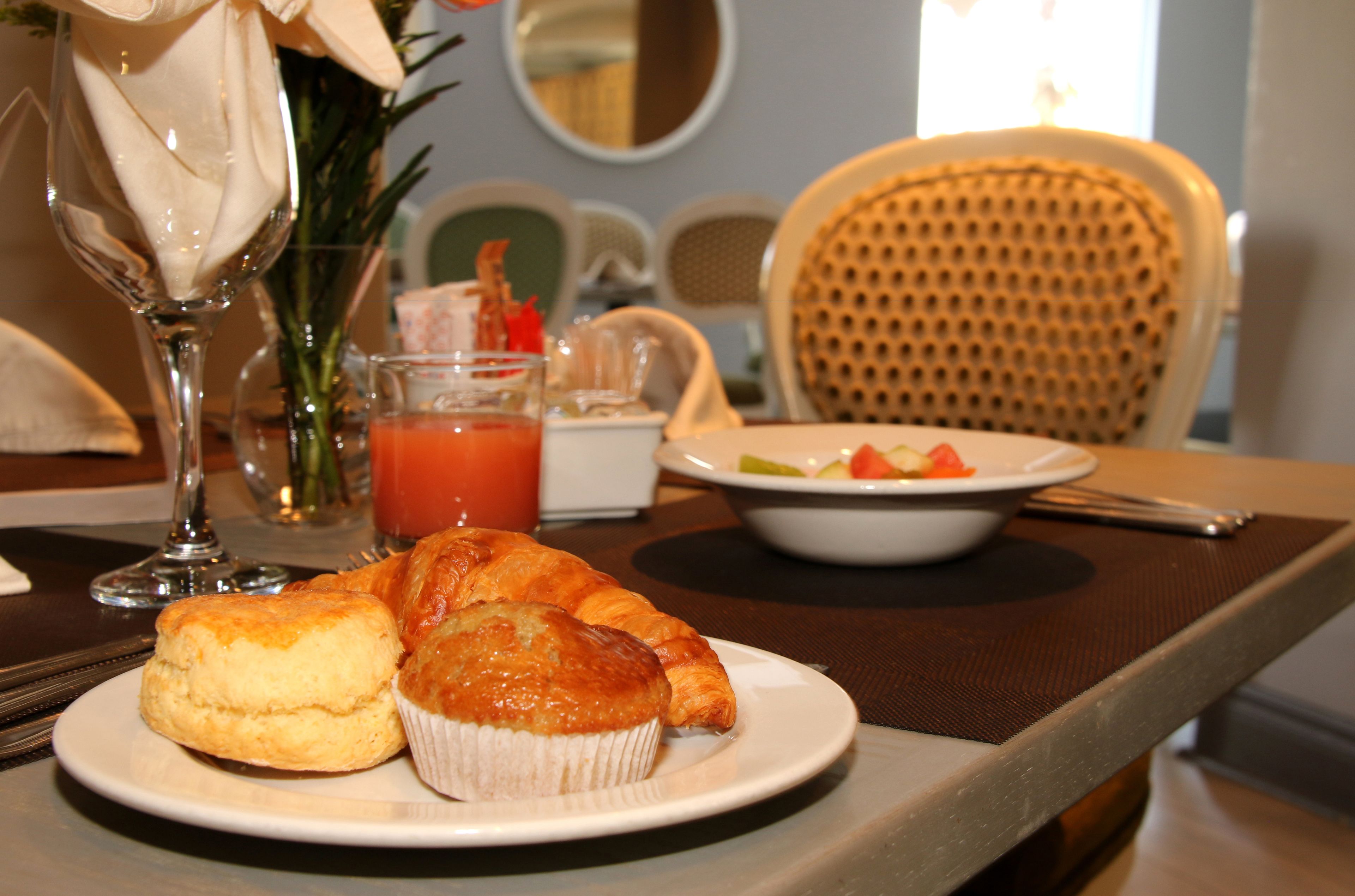 Un desayuno con una bandeja de muffins y croissants, un vaso de jugo y un bol de fruta sobre una mesa.
