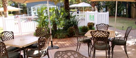 An outdoor patio area with tables and chairs under umbrellas, surrounded by greenery and a white fence.