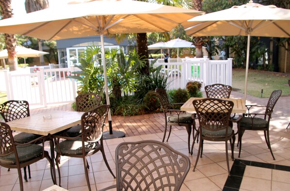 An outdoor patio area with tables and chairs under umbrellas, surrounded by greenery and a white fence.