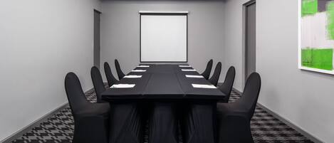 Un salón de conferencias con una larga mesa, sillas negras, una pantalla de proyector y alfombra estampada.